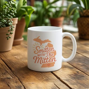 May include: White ceramic mug with the text "Smitten with The Mitten" in orange and peach tones, featuring a map of Michigan. The mug has a curved handle and is set on a wooden surface, with plants in the background.
