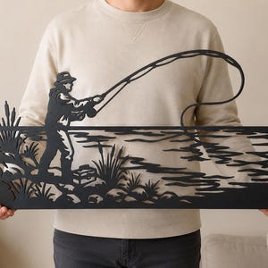 May include: A black metal wall art piece featuring a silhouette of a person fishing by a lake. The design includes a figure wearing a hat and holding a fishing rod, with cattails, a tackle box, and stylized water ripples depicted in the metal.