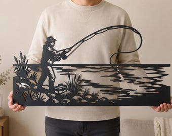 Puede incluir: Una pieza de arte mural de metal negro que presenta la silueta de una persona pescando junto a un lago. El diseño incluye una figura con sombrero que sostiene una caña de pescar, con espadañas, una caja de aparejos y ondas de agua estilizadas.