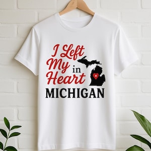 May include: White t-shirt with the text "I Left My Heart in MICHIGAN" in red and black. The design includes a black silhouette of the state of Michigan with a red heart. The shirt is hanging on a wooden hanger.