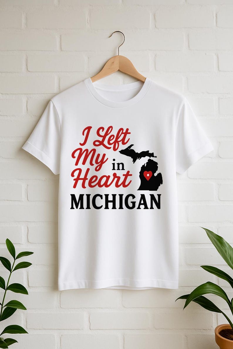 May include: White t-shirt with the text "I Left My Heart in MICHIGAN" in red and black. The design includes a black silhouette of the state of Michigan with a red heart. The shirt is hanging on a wooden hanger.