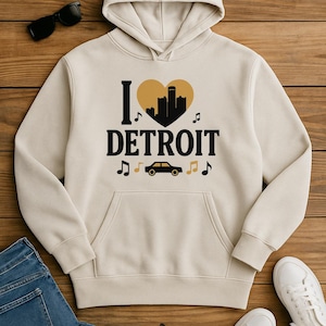May include: Cream hoodie with a front pocket, displaying "I ❤️ DETROIT" in black and gold. The design includes a heart-shaped cityscape, musical notes, and a car. Sunglasses, jeans, and white trainers are also visible.