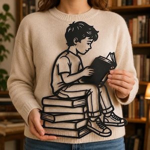 May include: A black metal wall art piece featuring a silhouette of a young person sitting on a stack of books while reading an open book. The design is rendered in a flat, cutout style with clean lines, showcasing the figure in a t-shirt and sneakers.