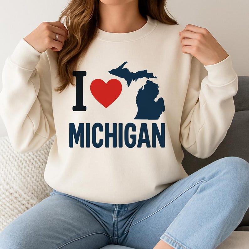 May include: A cream-coloured sweatshirt featuring the text "I ❤️ MICHIGAN". The "I" is black, the heart is red, and the state outline is navy blue. The word "MICHIGAN" is also navy blue. The sweatshirt has long sleeves and a crew neck.
