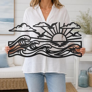 May include: A black metal wall art piece featuring a coastal scene with stylized ocean waves, a rising sun with radiating beams, clouds, and a bird in flight. The design is cut from a single flat sheet of metal with a matte black finish.