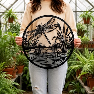 May include: A circular black metal wall art piece featuring a detailed cutout design of a dragonfly hovering over a pond with water lilies and cattails. The silhouette depicts a natural scene with aquatic plants and ripples on the water surface.