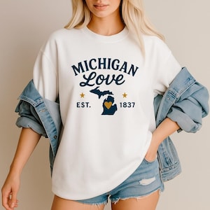 May include: White sweatshirt with the text "MICHIGAN Love" in navy blue, featuring a navy blue outline of the state of Michigan with a gold heart. The sweatshirt also includes the text "EST. 1837" and two gold stars.