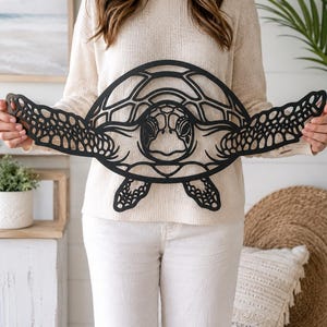 May include: A black, flat, laser-cut wall art piece shaped like a sea turtle. The design features intricate, open-work geometric patterns throughout the shell, head, and wide-spread flippers, creating a detailed silhouette of the marine animal.