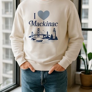 May include: A cream-coloured sweatshirt featuring the text "I ❤️ Mackinac" in blue, with a blue heart and illustrations of a bridge and lighthouse. The person is wearing blue jeans.