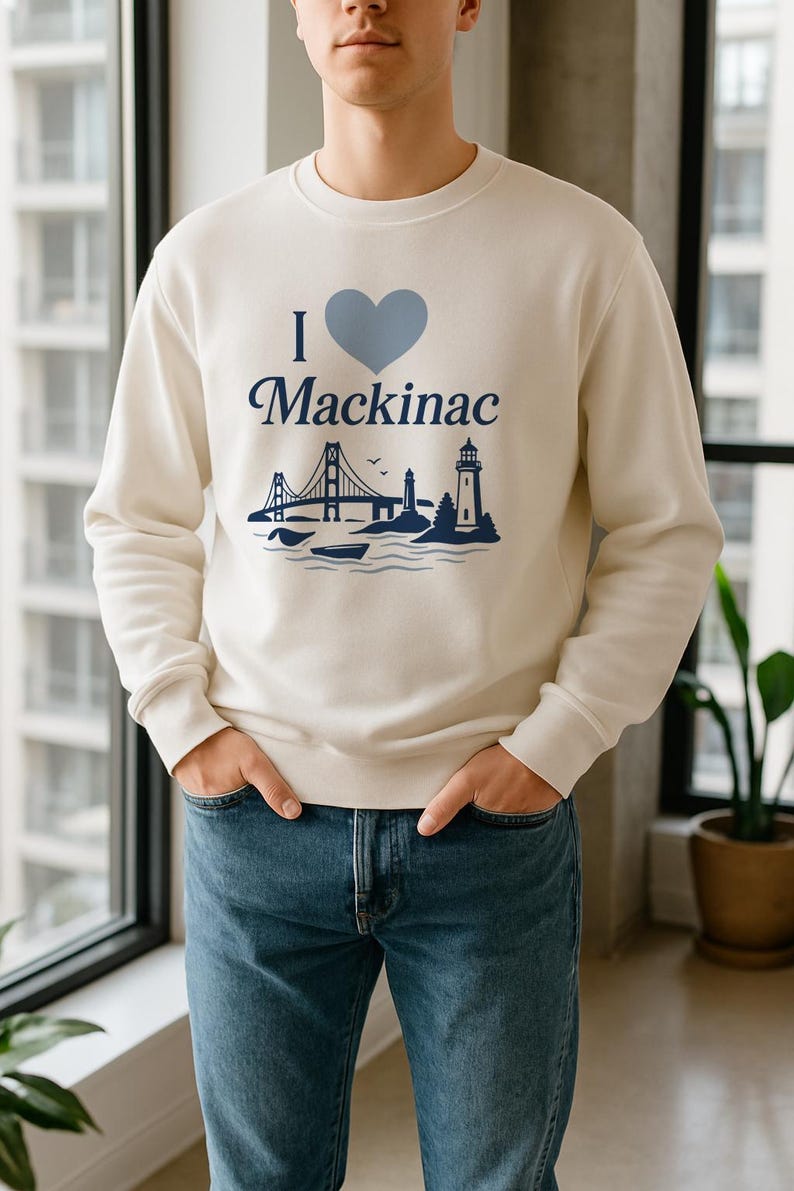 May include: A cream-coloured sweatshirt featuring the text "I ❤️ Mackinac" in blue, with a blue heart and illustrations of a bridge and lighthouse. The person is wearing blue jeans.