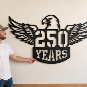 May include: A black metal wall art piece featuring an eagle with outstretched wings. The design includes the numbers "250" and the word "YEARS" cut out within the eagle's body. The art is mounted on a white wall.