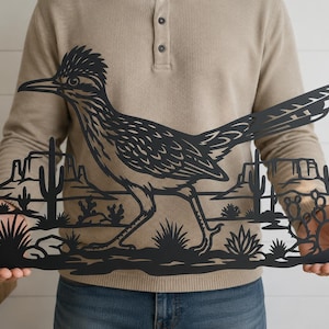 May include: A black metal wall art piece featuring a detailed silhouette of a roadrunner bird walking through a desert landscape. The design includes cutouts of saguaro cacti, prickly pear cacti, and distant mesas set against a flat horizon line.