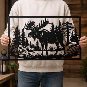 Può includere: Un'opera d'arte da parete rettangolare in metallo nero con una silhouette tagliata al laser di un alce in una foresta. Il design include pini dettagliati, felci e terreno roccioso all'interno di una cornice dalle linee pulite.