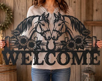Puede incluir: Un letrero de pared de metal negro con dos cabezas de caballo frente a frente, sobre un diseño de cerca de madera con girasoles y una herradura. La palabra WELCOME está recortada en letras mayúsculas grandes en la parte inferior.