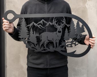 Puede incluir: Una pieza de arte mural de metal negro mate con forma de anzuelo grande. El recorte interior presenta una silueta detallada de un ciervo de pie, un denso bosque de pinos y una cadena montañosa al fondo, todo realizado en metal.