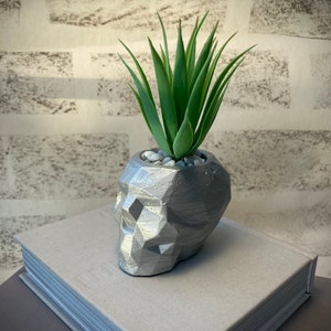 Maceta de hormigón color plata con forma de calavera y planta artificial.