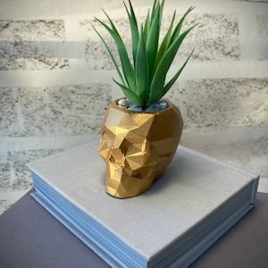 Macetero de calavera de hormigón dorado con planta artificial.