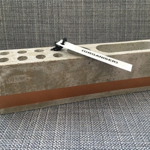 May include: A grey concrete desk organiser with a copper stripe and multiple compartments. The organiser has a label that says "(ORGANISER)".
