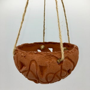 May include: A hanging terracotta planter with a textured surface and carved design. The planter is suspended by four natural fibre ropes. The interior of the planter is visible, and the background is plain white.