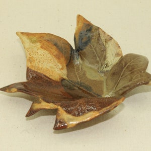 May include: A ceramic leaf-shaped dish with a naturalistic design. The dish features a blend of brown, tan, and green hues, with a glossy finish. The leaf has detailed veining and a slightly cupped shape, perfect for holding small items.
