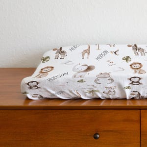May include: A white changing pad cover with a repeating pattern of safari animals in brown and tan. The word "HUDSON" is printed on the cover. The cover is on a wooden dresser.