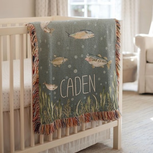 May include: A teal baby blanket with a fish design and the name "CADEN" in white lettering. The blanket features illustrations of various fish, bubbles, and aquatic plants. The edges are finished with a colorful fringe, and it is draped over a white crib.