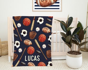 May include: A navy blue baby blanket with a sports theme. The blanket features a repeating pattern of baseballs, basketballs, footballs, bats, and gloves. The name "Lucas" is printed in white on the blanket.