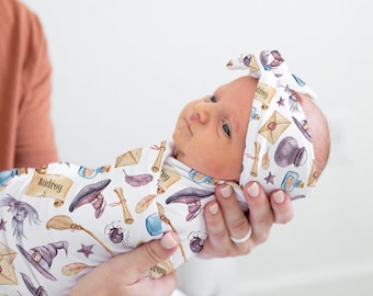 May include: A white swaddle blanket with a whimsical witch-themed print featuring stars, potion bottles, broomsticks, and a raven. The blanket is wrapped around a baby wearing a matching headband with the name "Audrey" on it.