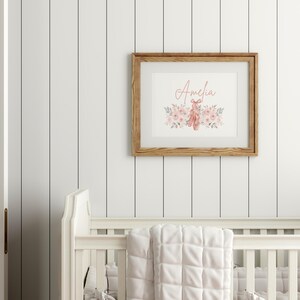 May include: A framed print with the name "Amelia" in a cursive font, surrounded by watercolor flowers and ballet shoes. The frame is made of light brown wood, and the print is displayed on a white wall.