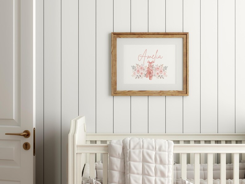 May include: A framed print with the name "Amelia" in a cursive font, surrounded by watercolor flowers and ballet shoes. The frame is made of light brown wood, and the print is displayed on a white wall.