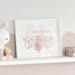 May include: A framed print with the name "Roselynn" in cursive script, featuring ballet shoes and floral accents in soft pink and gray tones. A white bunny figurine and decorative boxes are on a shelf.