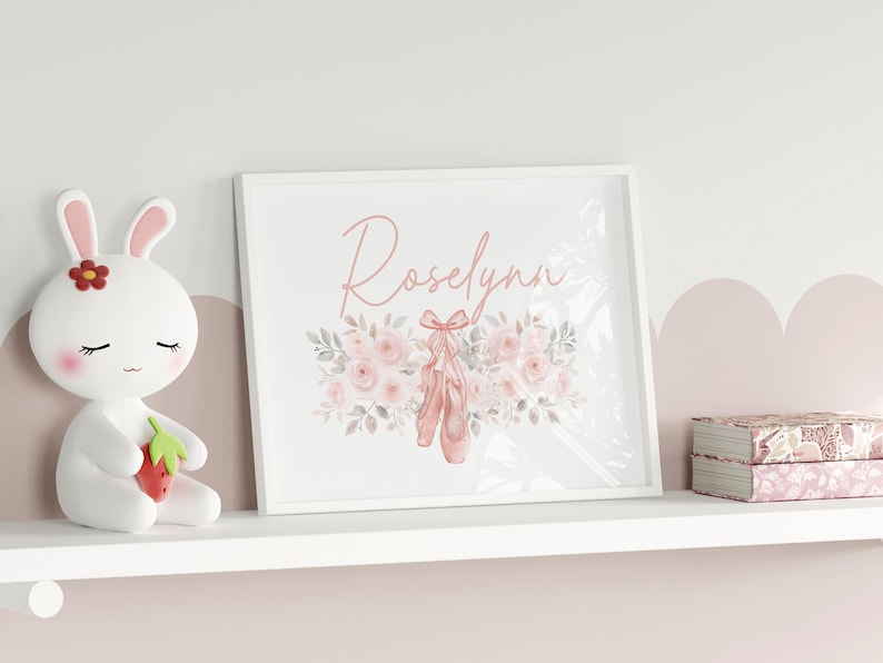 May include: A framed print with the name "Roselynn" in cursive script, featuring ballet shoes and floral accents in soft pink and gray tones. A white bunny figurine and decorative boxes are on a shelf.