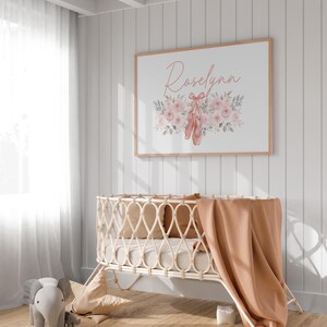 May include: A framed print with the name "Roselynn" in cursive script, surrounded by pink roses and ballet shoes. A light-colored woven baby crib with a peach blanket and pillows is in the foreground. A gray elephant toy is on the floor.