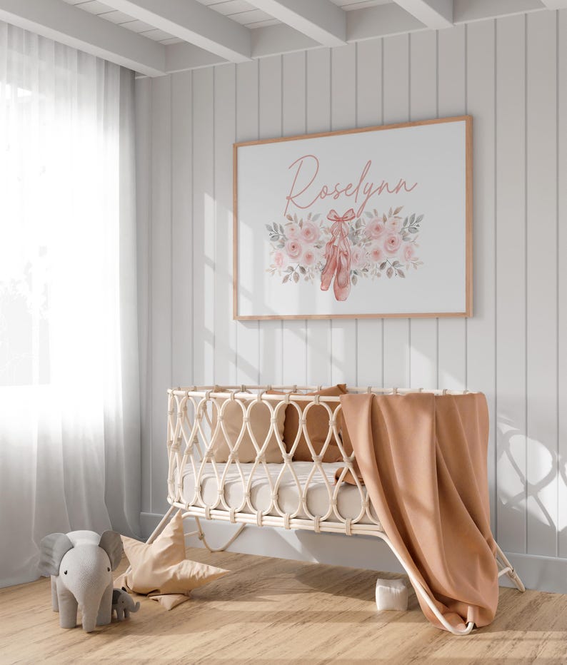 May include: A framed print with the name "Roselynn" in cursive script, surrounded by pink roses and ballet shoes. A light-colored woven baby crib with a peach blanket and pillows is in the foreground. A gray elephant toy is on the floor.