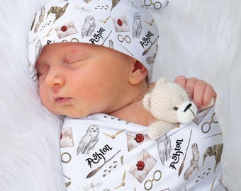 May include: A newborn baby wrapped in a white swaddle and matching hat, both patterned with owls, envelopes, glasses, and other magical symbols. A small, beige teddy bear is tucked in with the baby. The name "Ashton" is printed on the fabric.