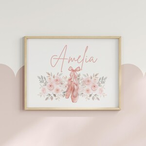May include: A framed print featuring the name "Amelia" in a delicate script above a watercolor illustration. The artwork showcases pink ballet shoes tied with a bow, surrounded by a floral arrangement of pink roses and gray leaves. The frame is a light wood color.