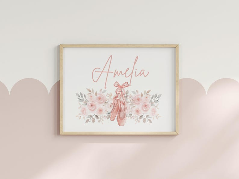 May include: A framed print featuring the name "Amelia" in a delicate script above a watercolor illustration. The artwork showcases pink ballet shoes tied with a bow, surrounded by a floral arrangement of pink roses and gray leaves. The frame is a light wood color.
