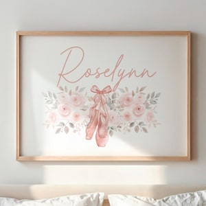 May include: A framed print featuring the name "Roselynn" in a soft pink script. Below the name is a floral arrangement with pink roses, gray leaves, and a pair of pink ballet shoes. The frame is a light wood color.