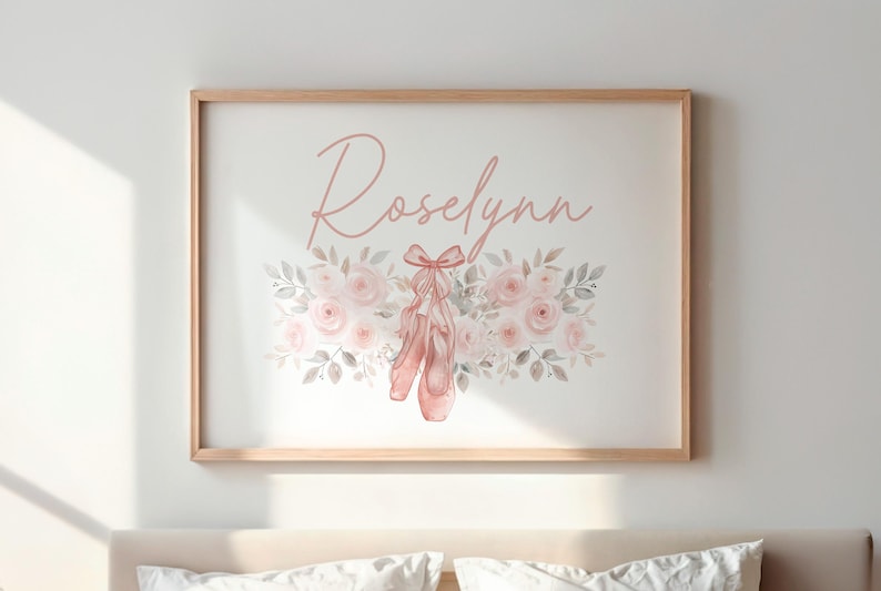 May include: A framed print featuring the name "Roselynn" in a soft pink script. Below the name is a floral arrangement with pink roses, gray leaves, and a pair of pink ballet shoes. The frame is a light wood color.