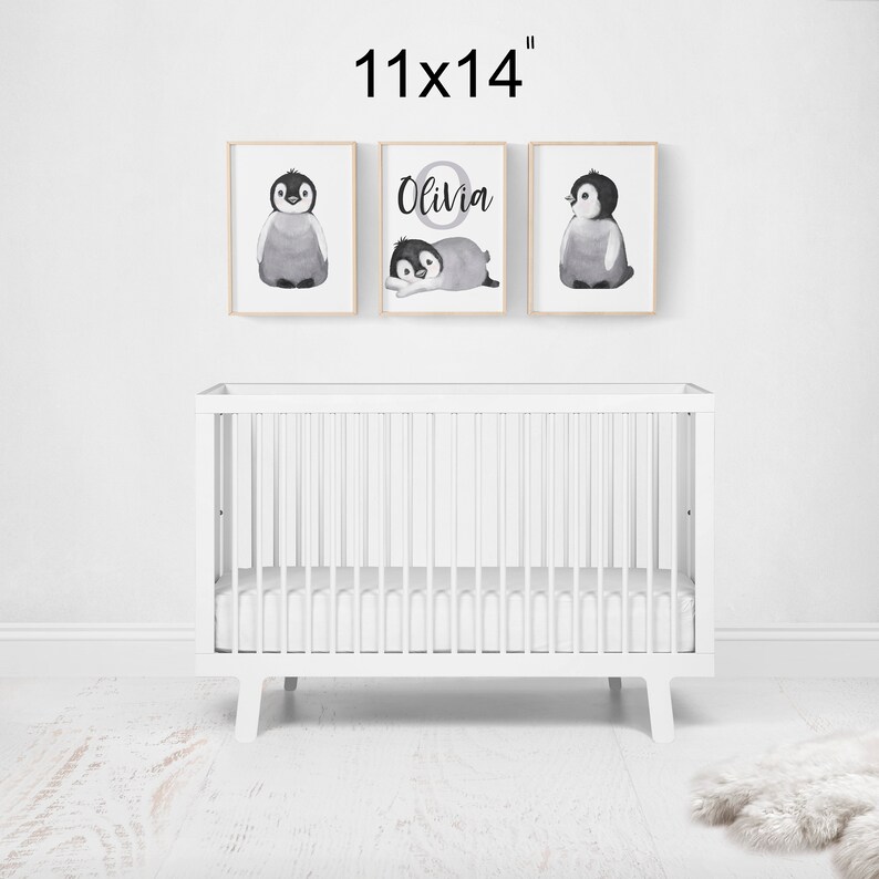 May include: Three framed prints with watercolor penguin illustrations. The center print features the name "Olivia" in a circle with a penguin inside. The prints are 11x14 inches and are displayed above a white crib.