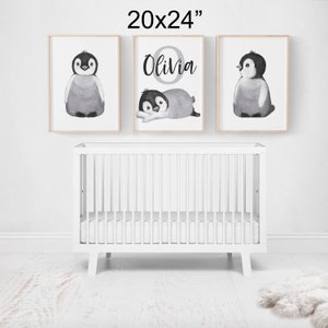 May include: Three black and white watercolor penguin illustrations in 20x24 inch frames. The center frame features the name "Olivia" in a gray circle. The frames are hung on a white wall above a white crib.