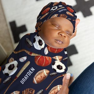 May include: A newborn baby wrapped in a navy blue swaddle and matching cap, both patterned with sports equipment illustrations. The pattern includes footballs, baseballs, basketballs, soccer balls, and hockey pucks. The name "BRAYDEN" is printed on the swaddle.