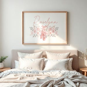 May include: A framed print with the name "Roselynn" in cursive script, surrounded by pink flowers, leaves, and ballet shoes. The artwork is displayed above a bed with white bedding and pillows. The frame is made of wood.