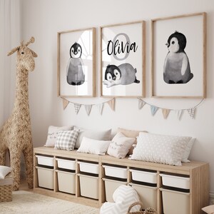 May include: Three framed prints with watercolor illustrations of penguins. The center print features the name "Olivia" in a circle with a penguin inside. The prints are hung on a white wall above a wooden shelf with storage bins.
