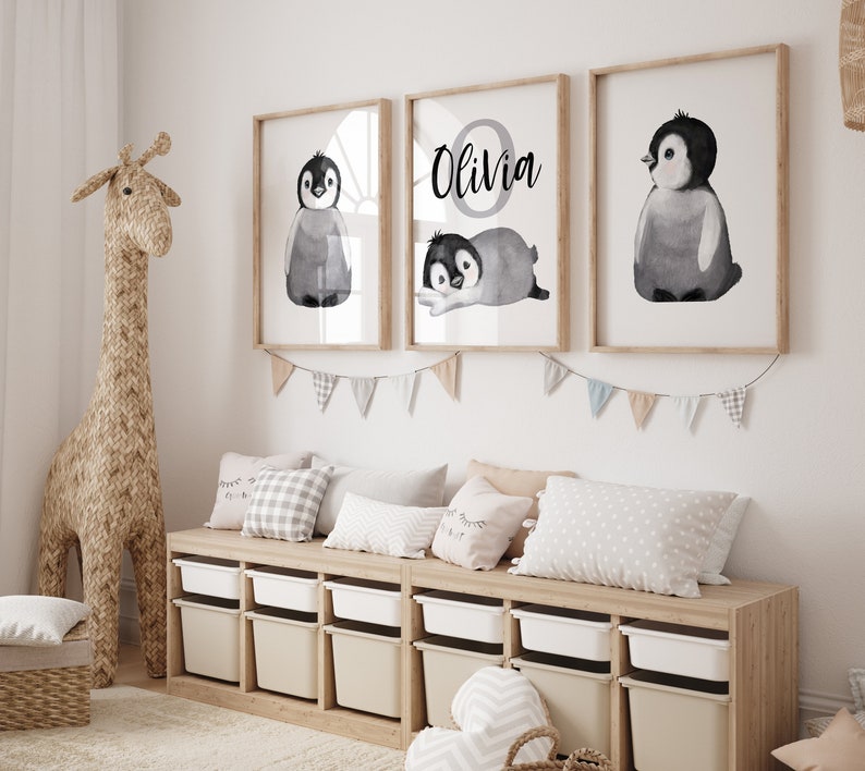 May include: Three framed prints with watercolor illustrations of penguins. The center print features the name "Olivia" in a circle with a penguin inside. The prints are hung on a white wall above a wooden shelf with storage bins.