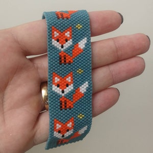 Puede incluir: Un brazalete de cuentas azul turquesa con un patrón repetido de caras de zorros naranjas y blancas. El brazalete está hecho con cuentas pequeñas y cuadradas.