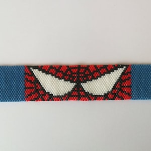 以下が含まれることがあります： スパイダーマンのデザインが施された赤、黒、白のビーズブレスレット。ブレスレットは小さな正方形のビーズで作られており、青い縁取りがあります。