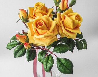 May include: A bouquet of yellow roses with green leaves and red buds in a clear glass vase with a burgundy ribbon.
