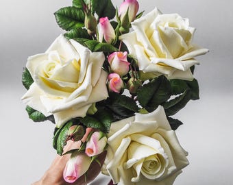 May include: A bouquet of white roses with pink buds, arranged with green leaves. The roses are held in a hand with a burgundy ribbon.