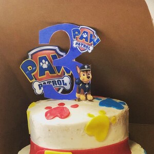 K&ouml;nnte beinhalten: Eine zweist&ouml;ckige wei&szlig;e Torte, verziert mit Pfotenabdr&uuml;cken, einem roten G&uuml;rtel und den Buchstaben "ALLA" in Blau, Gelb und Rot. Die Torte ist mit einer Paw Patrol Figur und einer blauen Zahl "3" mit den Worten "PAW PATROL" darauf verziert.